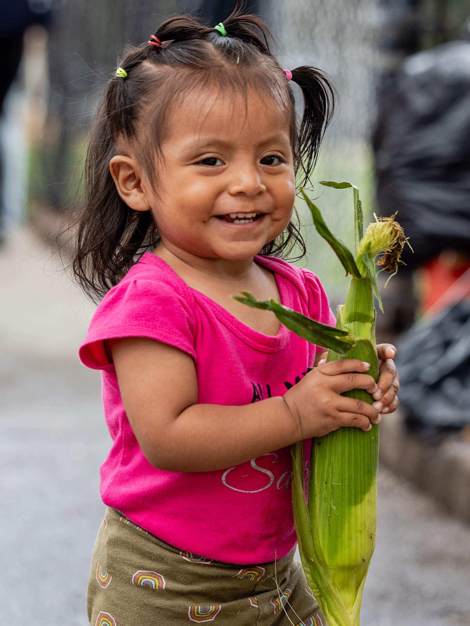 A young girl with pigtails smiles while holding a fresh ear of corn, standing outdoors in casual clothing.