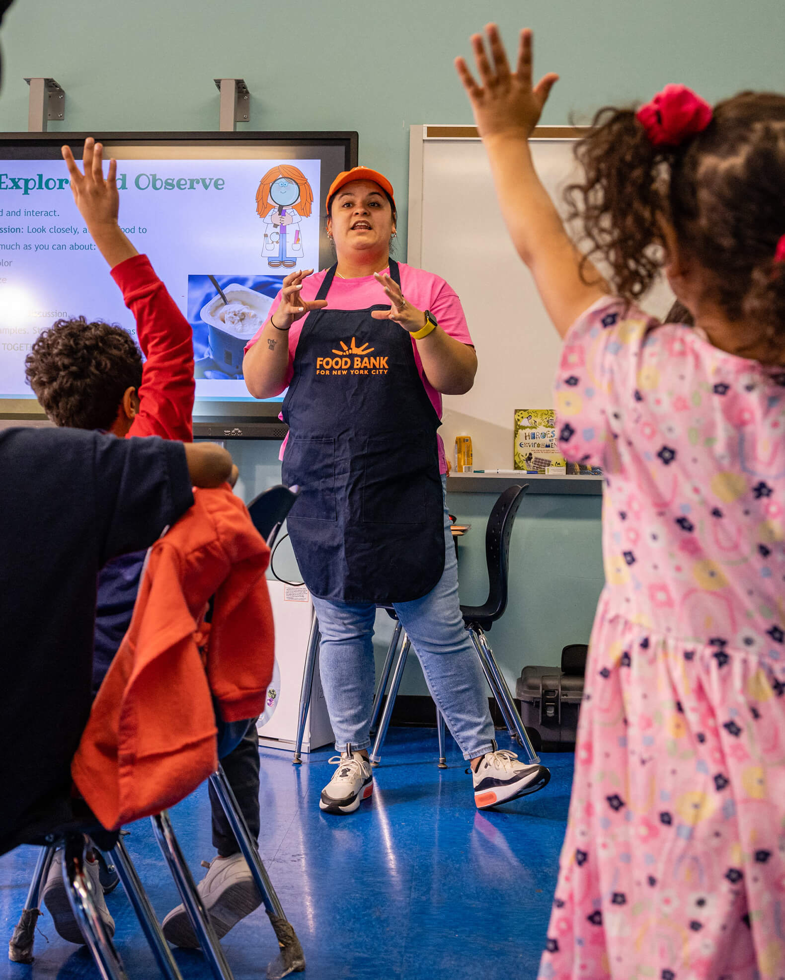 A woman in a Food Bank apron speaks to a classroom of children with raised hands, standing in front of a whiteboard and a presentation screen.